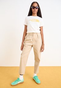 Beige byxor med hög midja, vit t-shirt med gul logotyp och mintgröna sneakers. Solglasögon med mörk toning, flätat hår.