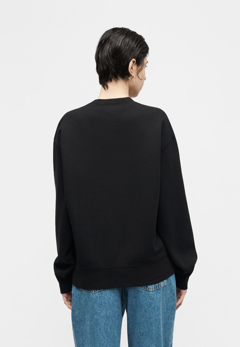 Persona con capelli corti e scuri, che indossa una felpa nera a maniche lunghe e jeans blu, vista di spalle su uno sfondo bianco uniforme.