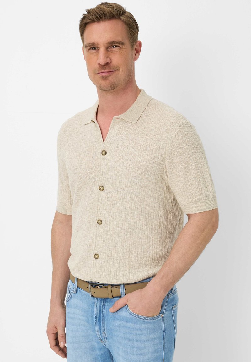 Mann mit kurzen hellbraunen Haaren, der ein beigefarbenes Kurzarmhemd mit Knöpfen und hellblaue Jeans mit einem hellbraunen Gürtel trägt.