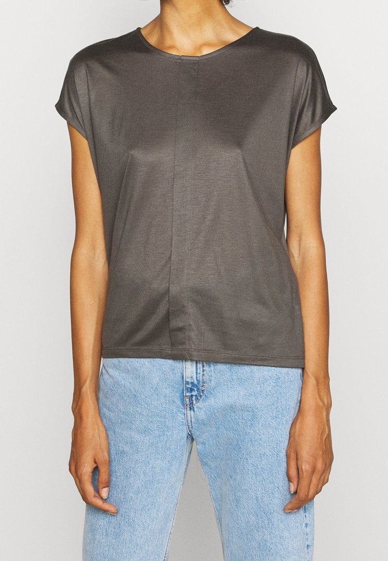 Femme portant un haut gris foncé à manches courtes avec un col rond et un jean en denim bleu clair, debout devant un fond uni.