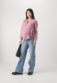 Rosa stickad omslagskofta med knyt detalj, i kombination med ljusblå utsvängda jeans och svarta ankelboots, hållande en svart handväska.