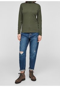 Olijfgroen langemouwen shirt met hoge hals; blauwe versleten jeans met gescheurde knie; bruine veterschoenen met een gestructureerde afwerking.