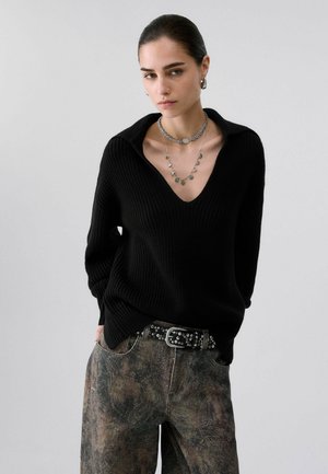 Femme aux cheveux tirés en arrière, portant un pull en maille noir, un pantalon à motifs, une ceinture cloutée, des boucles d'oreilles créoles et des colliers en argent superposés sur un fond uni.