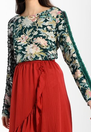 Kwiatowy top z długim rękawem w ciemnozielonym kolorze z różowymi i kremowymi kwiatami, w połączeniu z czerwoną, falbanianą spódnicą z zakładanym fasonem i teksturowaną tkaniną.