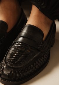 Mocasines negros tejidos de cuero, usados con pantalones negros, mostrando costuras detalladas y una banda superior de textura suave.