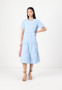 Vestido de algodão azul claro com mangas curtas, design em camadas, gola redonda e uma saia solta. Modelo segura uma bolsa rosa. Sandálias de tiras castanhas.