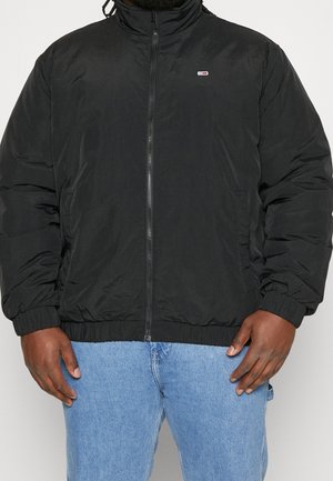 Hombre con chaqueta negra con cremallera y puños elásticos, y vaqueros azul claro, de pie frente a un fondo liso.