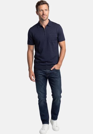 Mężczyzna ubrany w granatową koszulkę polo z krótkim rękawem i kieszenią na piersi, ciemnoniebieskie dżinsy oraz białe trampki, stojący z jedną ręką w kieszeni.
