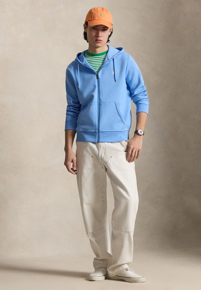 Noor mees oranži nokamütsiga, sinise lukuga kapuutsiga, rohelise triibulise särgiga, valgete pükste ja valgete tossudega, seisab beežis taustal.