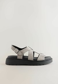 Sandale en daim gris avec design à lanières croisées, boucle à la cheville et semelle épaisse en caoutchouc noir. Dispose d'une semelle plate texturée et d'un bout ouvert.