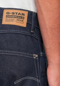 Dunkelblaue Jeans aus Denim mit einem braunen Lederpatch, das "G-STAR ORIGINALS RAW DENIM" trägt. Kontrastnähte und ein Detail der Gesäßtasche sind sichtbar.