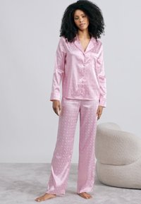 Rosa Satin-Pyjama-Set mit einem langärmeligen Hemd mit Knopfleiste und einer Hose mit dezentem Muster. Weiche Textur und lockere Passform.
