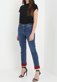 Blå denimjeans med rak passform, röd spetsdetalj vid kanten, i kombination med en svart t-shirt med kort ärm och svarta stilettklackar.
