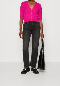 Cárdigan de punto fucsia con mangas cortas y patrón de diamantes, cierre de botones, combinado con pantalones vaqueros negros de cintura alta y zapatos negros.