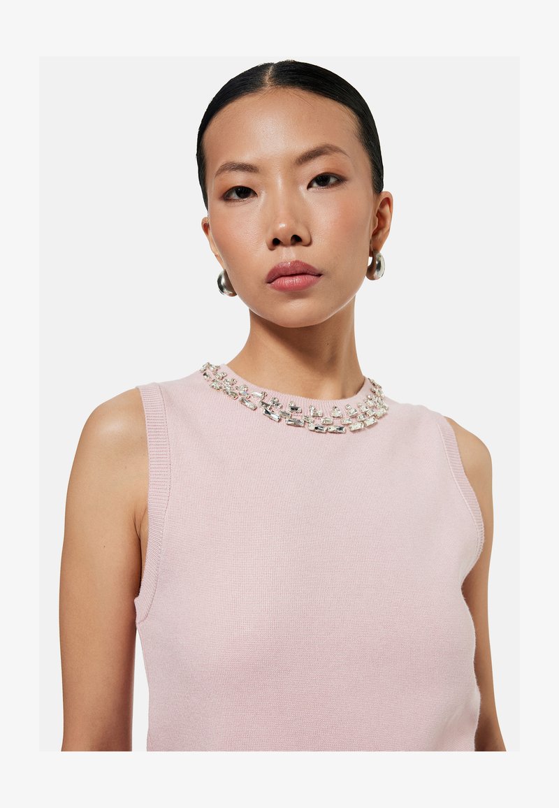 Young woman with sleek hair wearing silver hoop earrings and a sleeveless light pink top embellished with clear rectangular beads at the neckline.