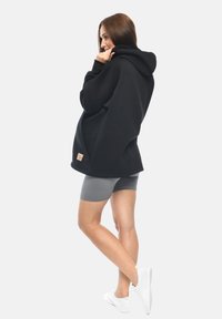 Felpa nera oversize con cappuccio e una piccola etichetta con logo. Il modello la abbina a pantaloni corti grigi e sneakers bianche. Texture liscia su tutta la superficie.