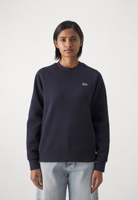 Marinblå hettegenser med rund hals, raglanermer og ribbede mansjetter. Har en liten grønn brodert logo på brystet.