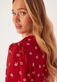 Blusa rossa con motivo floreale bianco, maniche a sbuffo e vestibilità rilassata. Tessuto liscio con una consistenza morbida. Vista ravvicinata di lato.
