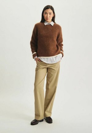 Pull en maille marron avec encolure arrondie, superposé à une chemise à col rayée blanche, associé à un pantalon beige ajusté et des chaussures foncées.