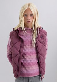 Niña rubia con el pelo largo, vestida con un suéter morado con estampado y una chaqueta sin mangas acolchada a juego, de pie frente a un fondo liso.