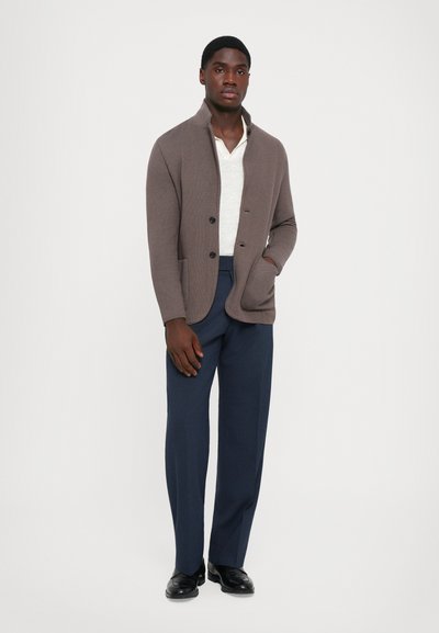 Man wearing brown buttoned cardigan over white shirt, navy trousers, and black shoes, standing against plain white background.