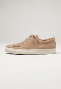 Beige suède sneaker met ronde neus, veters aan de voorkant, contrasterende off-white rubberen zool en subtiele stiksel details. Merkembleem aan de zijkant.