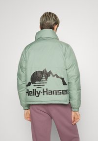 Vadderad grön jacka med hög krage och elastiska ärmslut. Har en svart grafik av berg och sol med texten "Helly-Hansen" på ryggen.