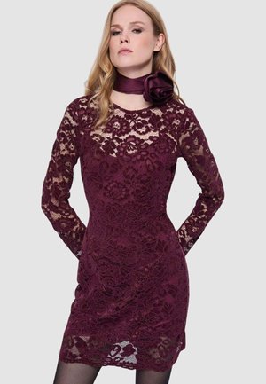 Donna che indossa un abito aderente a maniche lunghe in pizzo color bordeaux con motivi floreali e una sciarpa abbinata bordeaux con una grande rosellina.