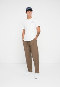 Hvit kortermet t-skjorte med blå grafikk, brune teksturerte bukser og hvite joggesko. Modellen har på seg en blå caps og står med hendene i lommene.