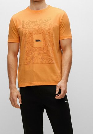 Mężczyzna w pomarańczowej koszulce z krótkim rękawem z geometrycznym wzorem tunelu i napisem "BOSS", w połączeniu z czarnymi spodniami z logo "BOSS".
