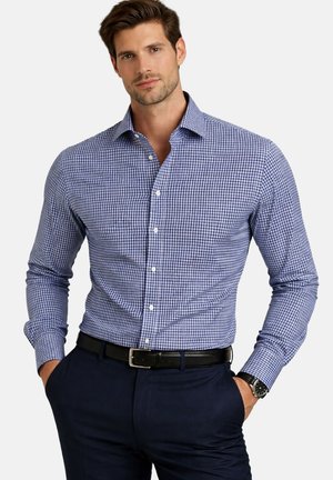 Homme portant une chemise à carreaux bleue et blanche boutonnée, un pantalon bleu foncé, une ceinture noire et une montre-bracelet, les mains dans les poches sur fond blanc.