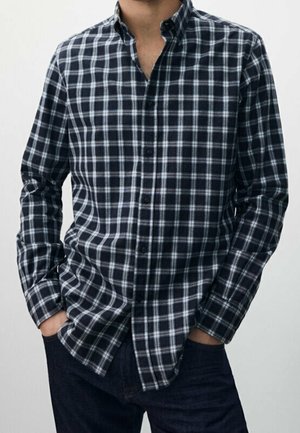 Mężczyzna ubrany w czarno-białą koszulę na guziki w kratkę z długimi rękawami oraz ciemne spodnie, z rękami swobodnie schowanymi w kieszeniach.