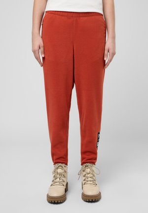 Rote Jogginghosen mit strukturiertem Finish, elastischem Bund, Seitentaschen und einem Logopatch am linken Bein. Kombiniert mit beige Wanderschuhen.