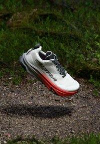 Hvit løpesko med teksturert overdel, svarte lisser og en rød-oransje såle. Har en polstret design og Vibram yttersåle for grep.