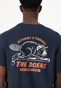 Camiseta azul marino con un gráfico de un castor sosteniendo un martillo, texto "ELEMENT X TIMBER! ¡LOS HACEDORES CONSTRUYEN ALGO" en naranja.