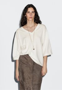 Light beige V-neck sweater with short sleeves, made of a soft material, paired with a dark brown suede skirt featuring subtle stitching details.