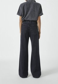 Henkilö, jolla on päällään haalistunut musta lyhythihainen paita, joka on sisätetty korkeavyötäröisiin leveälahkeisiin mustiin farkkuihin, seisoo vaaleaa taustaa vasten.