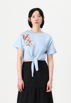 Maglietta corta azzurro chiaro con ricami che raffigurano fiori, maniche corte e dettaglio con nodo sul davanti, abbinata a una gonna nera a più strati.