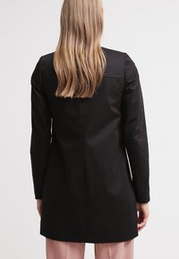 Nainen, jolla on pitkät vaaleat hiukset, pukeutuneena istuvaan mustaan bleiseriin ja vaaleanpunaiseen housupukuun, seisoo selkä kameraa kohti valkoista taustaa vasten.