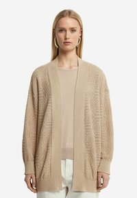 Beige stickad cardigan med ett texturerat geometriskt mönster, öppen framsida, långa ärmar och en avslappnad passform. Bärs över en matchande topp.