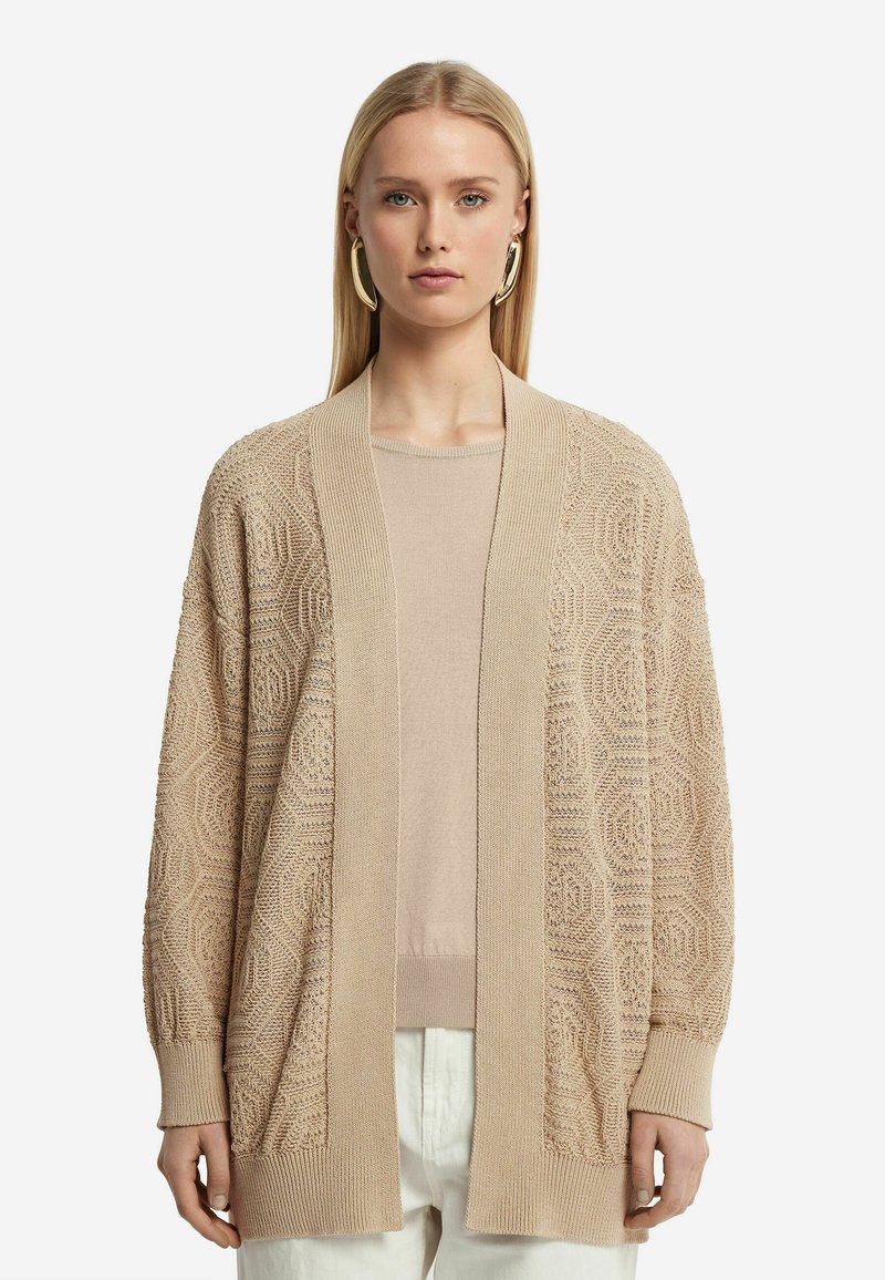 Beige stickad cardigan med ett texturerat geometriskt mönster, öppen framsida, långa ärmar och en avslappnad passform. Bärs över en matchande topp.