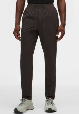 Mann trägt dunkelbraune Slim-Fit-Hose mit Tunnelzugbund und weißen Schnürsneakern vor einfarbigem Hintergrund.