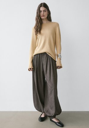 Frau mit langen braunen Haaren, die einen beigen Pullover, weite braune Nadelstreifenhosen, schwarze Ballerinas und ein silbernes Manschettenarmband am rechten Handgelenk trägt.