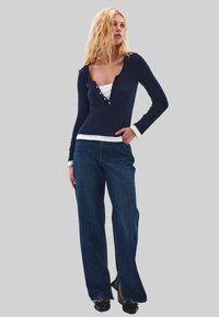 Blonde Frau in navyblauem Langarmshirt, das über einem weißen Oberteil getragen wird, mit blauen weiten Jeans und schwarzen Schuhen, steht mit einer Hand in der Tasche.