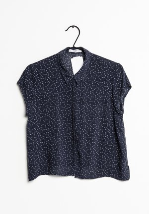 Blusa blu navy a maniche corte con colletto, caratterizzata da un modello geometrico bianco ripetitivo su tutto il tessuto. Esposta su un appendiabiti nero.