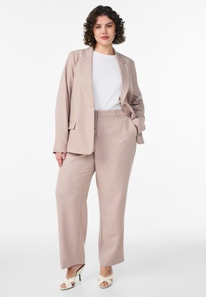 Kobieta o kręconych ciemnych włosach, ubrana w beżowy garnitur z szerokimi spodniami, białą bluzkę i białe sandały na obcasie z odkrytymi palcami, stojąca z jedną ręką w kieszeni.