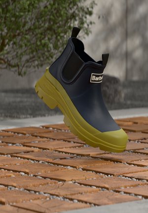 Тъмносиня и жълта гумена глезенна обувка с лого Barbour, показана да плава над дървено тухлено настилка навън близо до зеленина.