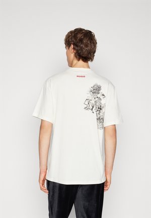 Jeune homme aux cheveux bruns courts portant un T-shirt blanc avec un petit logo rouge "HUGO" et un croquis en noir et blanc de la nature dans le dos.