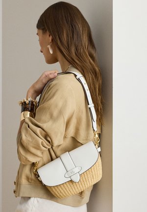 Kvinde med langt brunt hår iført beige jakke og armbånd, som holder en vævet taske med hvid læderklap og justerbar skulderrem.