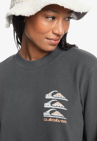 Grijze sweatshirt met ronde hals bevat het Quiksilver-logo in wit en oranje. Model draagt een fuzzy crème emmerhoed.
